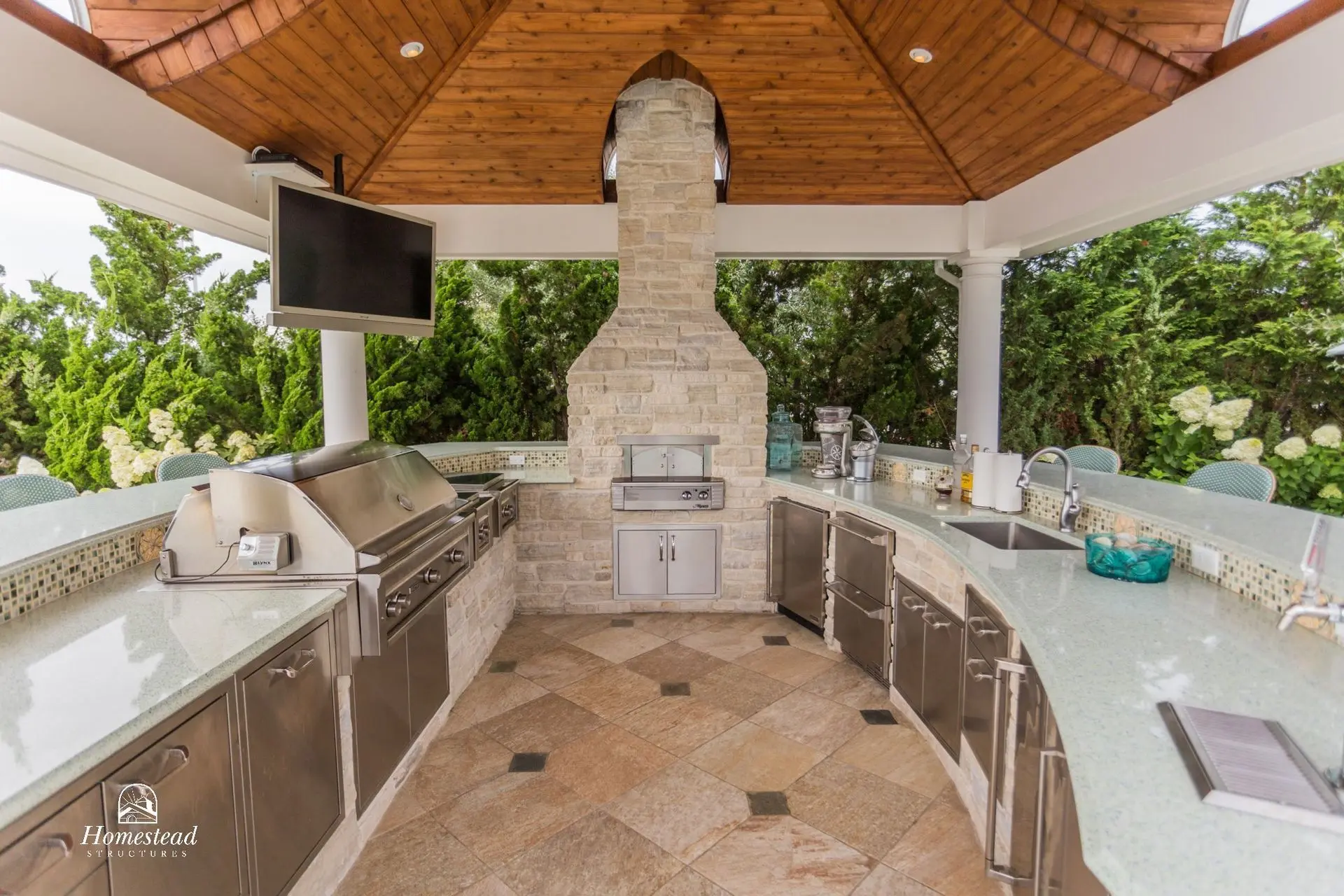 Covered outdoor kitchen with a stainless grill, stone chimney, and wood ceiling