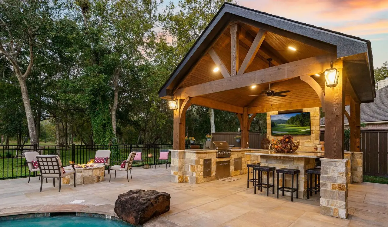 Gable pavilion with an outdoor kitchen, bar seating, and lighting beside a pool at dusk