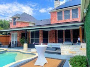 Backyard with a pool, patio, and brick house under a blue sky; a white table in the foreground.