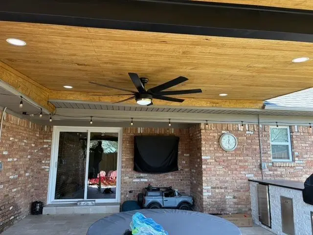 Stained wood patio ceiling with a large fan and string lights on a brick home