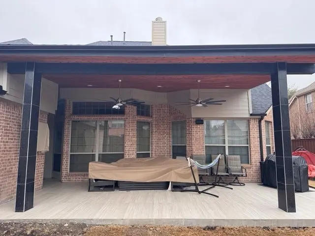 Patio cover with black posts over a wood deck behind a brick home